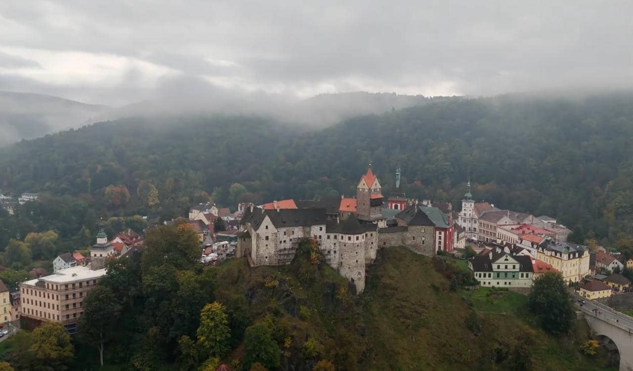 Luftaufnahme der Burg Loket über der Stadt, im Hintergrund bewaldete Hügel im Nebel