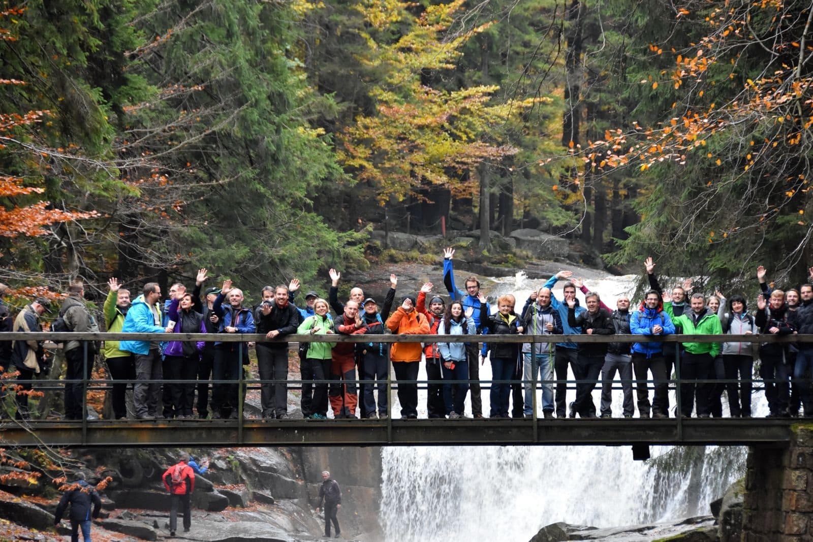 Gruppe in Outdoor-Kleidung winkt auf einer Brücke über einem Wasserfall