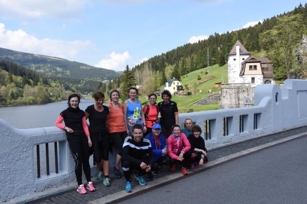 Laufteam in bunten Sportoutfits posiert auf einer Talsperre mit Blick auf See und Schloss
